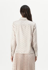 Blouse col montant avec un motif en crème, beige et rose pâle, dotée de manches longues et d'une coupe droite, portée avec une jupe plissée.