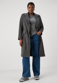 Pasi sivo prugast dolgo plašč z reverji, žepi in sproščenim krojem, kombiniran s sivim topom in širokimi modrimi kavbojkami, dopolnjenimi s srebrnimi supergami.