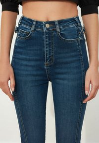 Jeans de mezclilla azul de cintura alta con un corte ajustado, ligero desgaste y detalles metálicos en la cinturilla y los bolsillos.