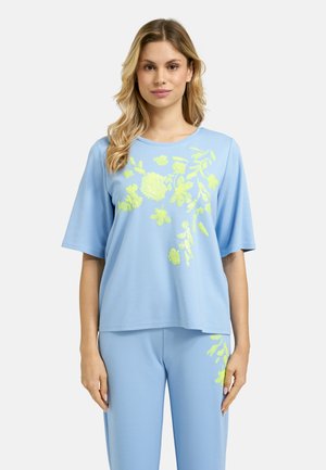 Woman wearing light blue loose-fit top and pants with yellow floral embroidery, standing against a plain light background.