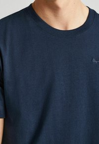 Hombre vistiendo una camiseta de cuello redondo azul marino con un pequeño logotipo bordado discreto en el lado izquierdo del pecho.