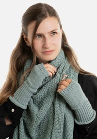 Jonge vrouw met lang haar past een gebreide lichtblauwe sjaal aan, met bijpassende vingerloze handschoenen en een zwarte jas.