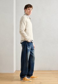 Beige hosszúujjú pulóver, alatta kék kockás ing, sötétkék farmernadrág és világosbarna hálós sportcipő szürke részletekkel.