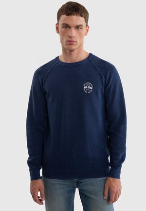 Granatowy sweter dzianinowy z teksturowanym wzorem, ściągacze przy mankietach i na dole, z białym logo na lewej piersi. Luźny krój, okrągły dekolt.