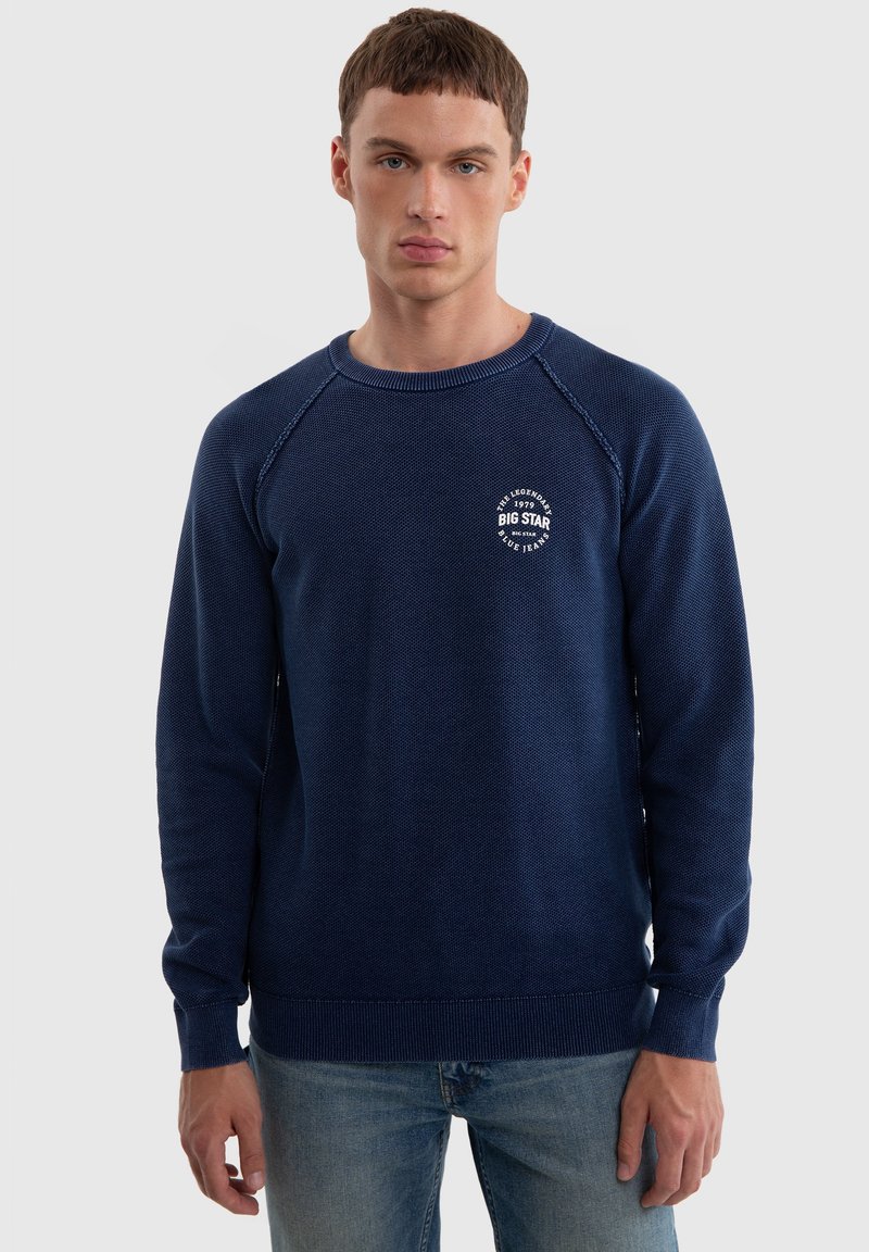 Námořnický modrý pletený svetr s texturovaným vzorem, žebrovanými manžetami a lemem, s bílým logem na levé hrudi. Pohodlný střih, kulatý výstřih.