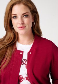 Veste zippée bordeaux avec boutons-pression, associée à un t-shirt graphique blanc. Présente une texture lisse et une coupe décontractée. Boucles d'oreilles en argent en forme de anneau.
