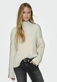 Maglione a coste crema con collo alto e dettagli a righe nere lungo le maniche, abbinato a jeans scuri e una cintura nera.