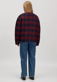 Rode en marineblauwe geruite oversized blouse met brede mouwen, gedragen met losse blauwe jeans en zwarte schoenen. De blouse heeft een boxy snit en een zachte textuur.