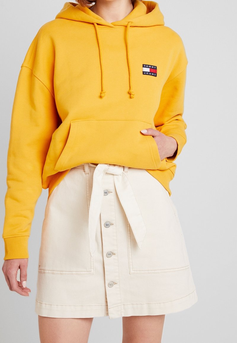 Sweat à capuche jaune avec cordons et poche avant, associé à une jupe en denim crème avec taille à nouer et fermeture à boutons.