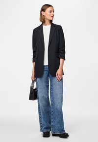 Blazer preto ajustado sobre uma t-shirt branca, combinado com jeans azuis de perna larga. Os acessórios incluem uma pequena mala preta e sapatos pretos robustos.