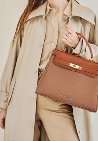 Sac à main structuré marron avec un dessus en cuir lisse et des accessoires en or, tenu par une personne portant un manteau et une chemise beiges.