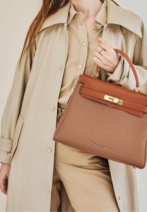 Hnědá strukturovaná kabelka s hladkým koženým vrškem a zlatými detaily, kterou drží osoba v béžovém kabátě a košili.