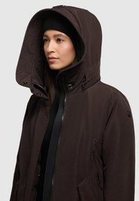 Chaqueta aislante marrón con una gran capucha, cierre delantero con cremallera y bolsillos laterales. Textura suave y características ajustables para un ajuste ceñido.