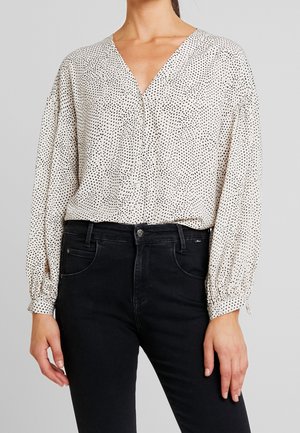 Femme portant une blouse blanche à manches longues avec de petits pois noirs rentrée dans un jean noir taille haute, debout devant un fond clair.