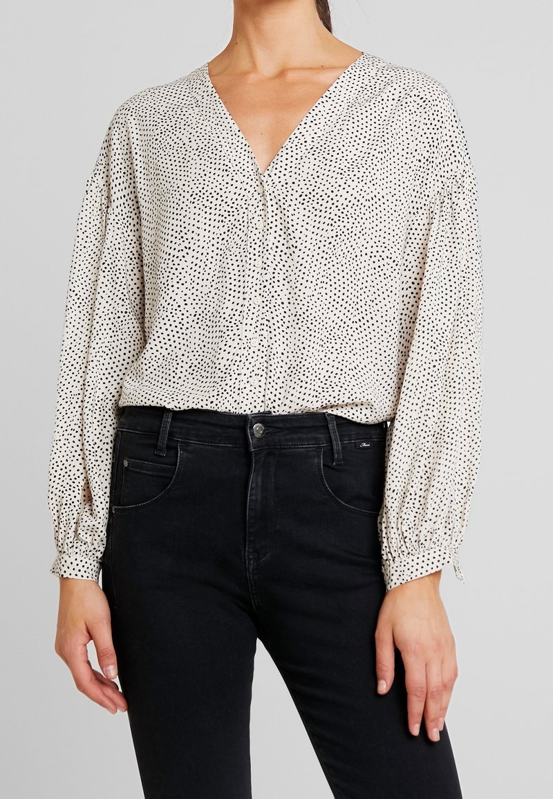 Femme portant une blouse blanche à manches longues avec de petits pois noirs rentrée dans un jean noir taille haute, debout devant un fond clair.