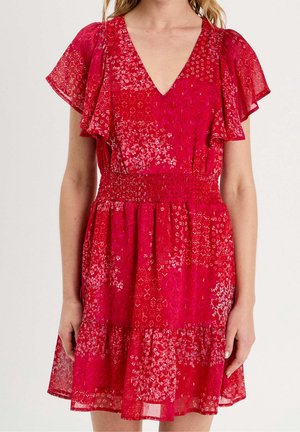 Kobieta w czerwonej sukience w kwiatowe wzory z krótkimi rękawami, ściąganym pasem, dekoltem w serek i falbaną na dole, widoczna od ramion do połowy uda.