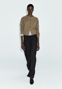 Beige gebreide cardigan met ronde halslijn, knoopsluiting en geribbelde manchetten, gecombineerd met zwarte wijde broek en zwarte puntige schoenen.