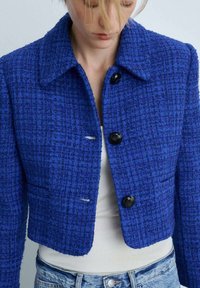 Modra tweed jakna s ovratnikom, ki ima teksturirano tkanino, štiri črne gumbe in dva stranska žepa. Skrajšan dizajn nad pasom.