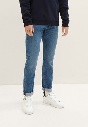 Vaqueros de mezclilla azul con corte recto, remangados en los tobillos. Combinados con zapatillas blancas con detalles negros. Diseño simple y casual.