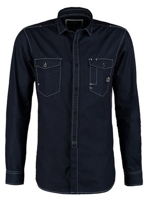 Mann trägt dunkelblaues Hemd, ausgewaschene schwarze Jeans, schwarze Schuhe und braunes Leder-Schulterholster und geht vorwärts vor einfarbigem Hintergrund.