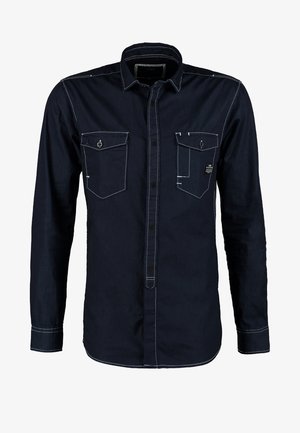 Hombre con camisa azul marino oscura, vaqueros negros desteñidos, zapatos negros y funda de hombro de cuero marrón caminando hacia adelante sobre un fondo liso.