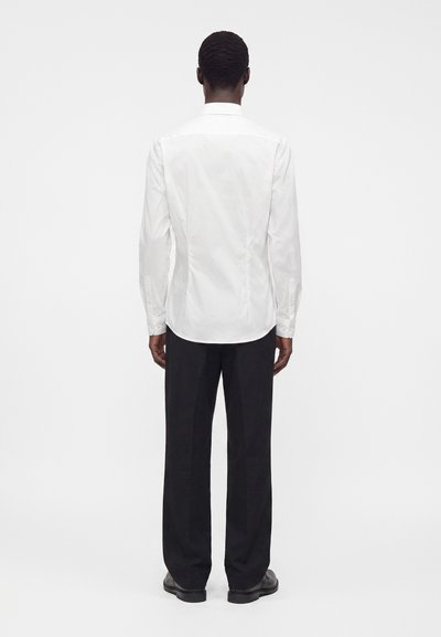 Uomo in piedi di spalle, che indossa una camicia bianca a maniche lunghe, pantaloni neri e scarpe nere su uno sfondo bianco uniforme.