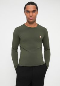 Bărbat îmbrăcat cu o cămașă verde măsliniu cu mâneci lungi și un mic logo roșu și alb pe piept, pantaloni negri, stând cu mâinile în buzunare pe un fundal alb.