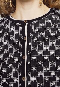 Cardigan in maglia nera con un motivo geometrico bianco, evidenziato da bottoni dorati e un bordo bianco a contrasto lungo il collo.