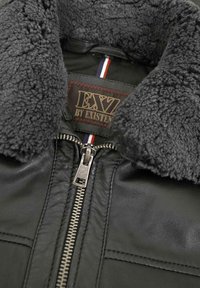 Schwarze Lederjacke mit einem flauschigen grauen Schaffellkragen, silbernem Reißverschluss und einem Stoffetikett mit rot, weiß und blauen Streifen am Kragen.