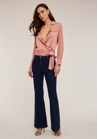 Blusa avvolgente in satin rosa con maniche lunghe, scollatura a V profonda e dettagli con fiocco, abbinata a jeans svasati blu scuro e sandali con cinturino.