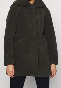 Abrigo de forro polar texturizado en gris oscuro, con capucha, bolsillos frontales y corte recto. Material suave y esponjoso con detalles de costura sutiles.