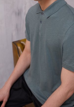 Osoba ubrana w szarozieloną koszulkę polo z krótkim rękawem i guzikami, siedząca na brązowym skórzanym fotelu, zdjęcie odcięte powyżej szyi.