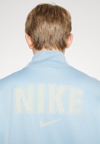 Felpa azzurro chiaro con collo alto e grande logo "NIKE" color crema sul retro, con un piccolo dettaglio del logo Nike swoosh.