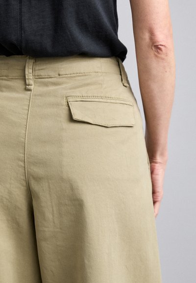 Beige trousers with a smooth texture, featuring a flat back pocket and a relaxed fit, paired with a black top, viewed from the back.