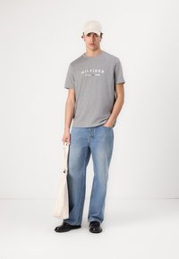 Siva pamučna majica s logom "HILFIGER", plave traperice, crne cipele i bež kapa. Drži bež torbu. Jednostavan, ležeran outfit.