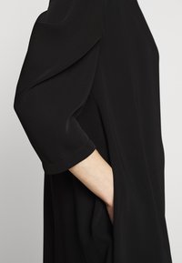 Svart blus med trekvartsärmar i ett mjukt, flödande tyg. Designen inkluderar en sidficka och subtila sömdetaljer.