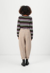 Personne aux cheveux bouclés portant une chemise à manches longues rayée, un pantalon beige en velours côtelé et des bottes noires, debout de dos sur un fond blanc.