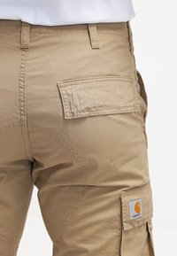 Beiga cargobyxor med en texturerad yta, bakficka med lock och synliga sömnadsdetaljer. Carhartt-logotyp på fickan.