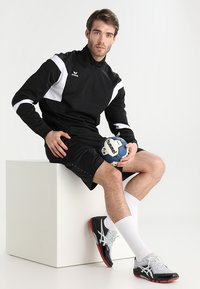 Schwarze Sportjacke mit weißen Akzenten, schwarze Shorts und graue Sportschuhe. Modell hält einen blauen Handball und sitzt auf einem weißen Block.