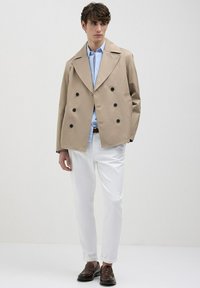 Giovane uomo in giacca doppiopetto beige, camicia azzurro chiaro, pantaloni bianchi e mocassini marrone scuro, in piedi davanti a uno sfondo bianco semplice.