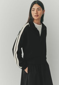 Veste zippée noire avec des rayures blanches sur les côtés, dotée d'un col montant et de poignets élastiques. Tissu doux avec une texture lisse.