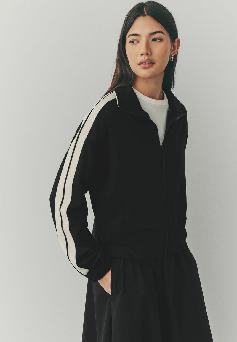 Veste zippée noire avec des rayures blanches sur les côtés, dotée d'un col montant et de poignets élastiques. Tissu doux avec une texture lisse.