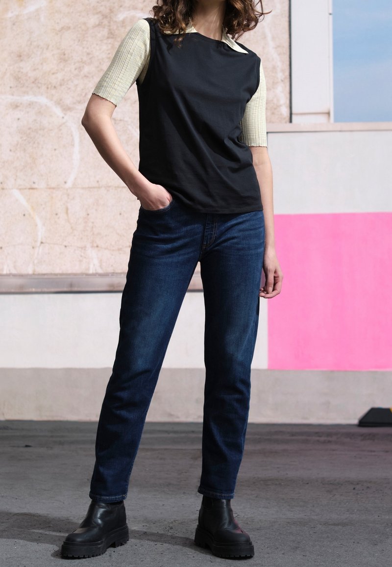 Černé tričko s krátkým rukávem a kontrastním prošívaným světle zeleným akcentem, spárované s tmavě modrými džíny a černými kotníkovými botami s hrubou podrážkou.