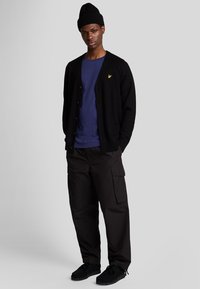 Cardigan nero sopra una t-shirt blu, pantaloni cargo neri e scarpe nere. Logo giallo ben visibile sul cardigan. Look casual, materiali texture.