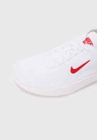 Hvid sneakers med overdel i tekstur, rød Nike swoosh-logo, flade snørebånd og en hvid gummisål med mønstret sål.