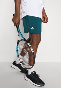 Teal spordilühikesed püksid valge Adidas logo, mustad tennised, valged sokid ja tennisereket sinise ja musta raamiga. 