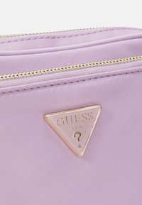 Lila nylonväska med en guldlås och en triangulär guldlogotavla som har "GUESS U.S.A." och en frågeteckensdetalj.