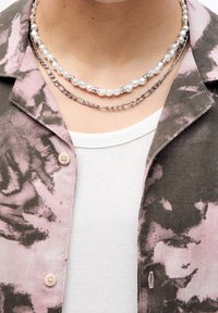 Collane a tre strati: una con perle bianche, una con dettagli in argento e una terza in catena argentata. Indossate sopra una maglietta bianca a coste e un giubbotto a fantasia.