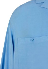 Chemise bleu clair à manches longues avec une poche avant boutonnée et un col pointu sur la partie supérieure gauche.