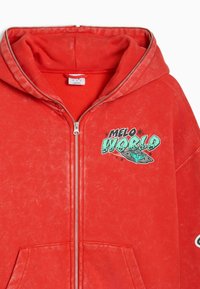 Punainen vetoketjullinen huppari etutaskuilla, teksti "MELO WORLD" ja lentävän lautasen kuva vasemmassa rinnassa.